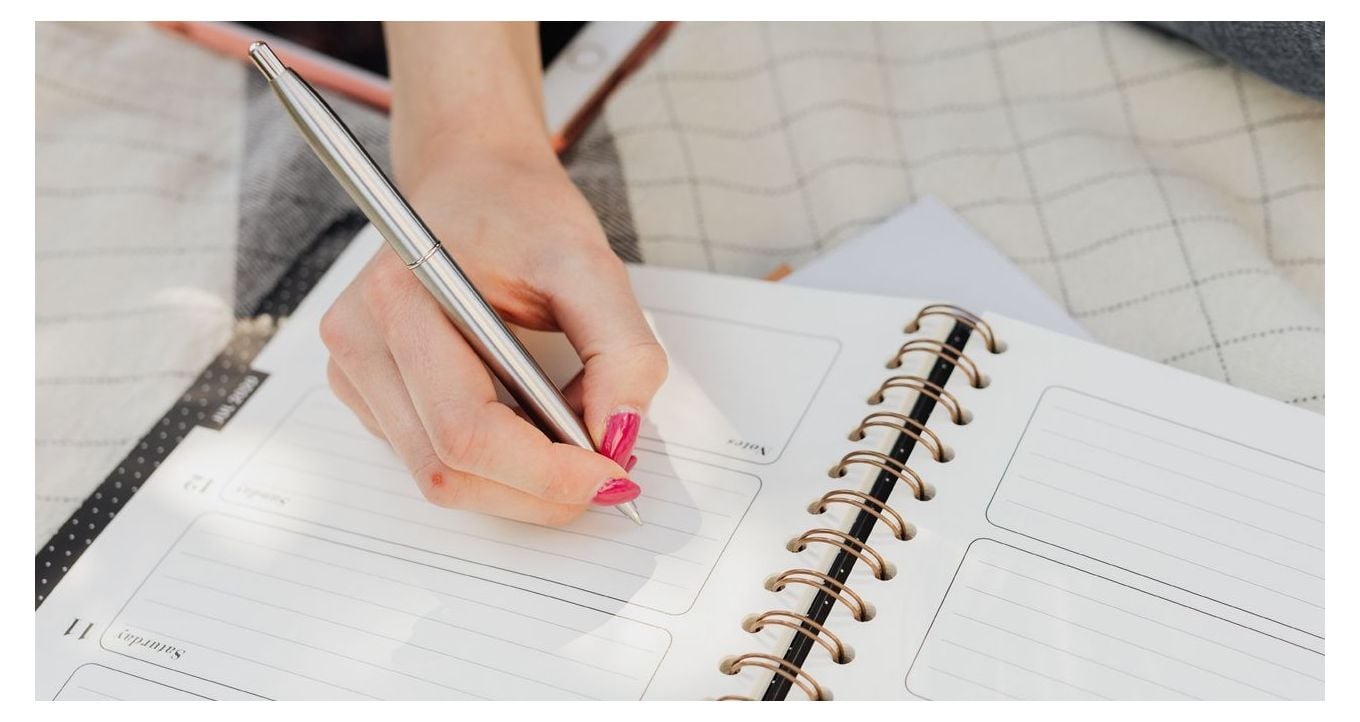 Hand holding a pen and writing on a page of a wire bound planner.