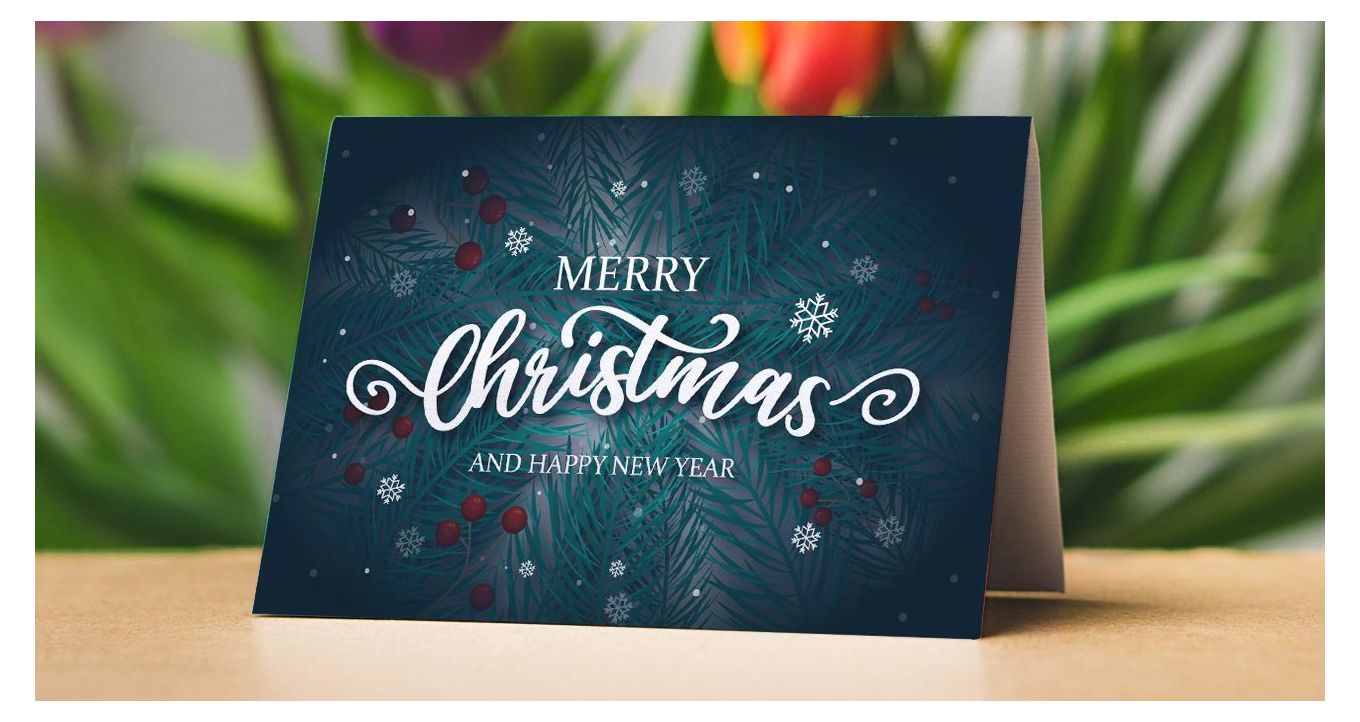 Front side of a Christmas-themed greeting card on a wooden desk and a flower arrangement behind it.