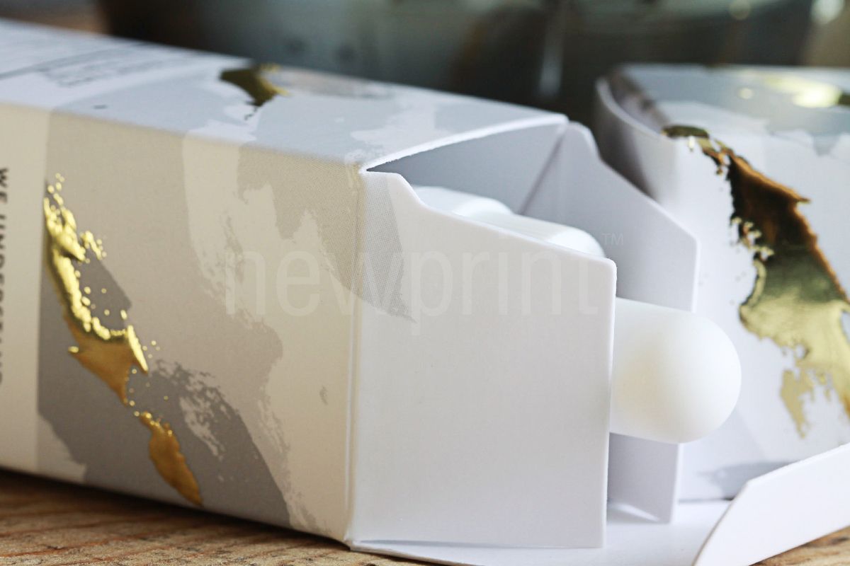 What is custom packaging - A close up of two small packaging boxes for CBD oil showing the structure of the white paperboard the box is made of, with gray and gold printing on the box.