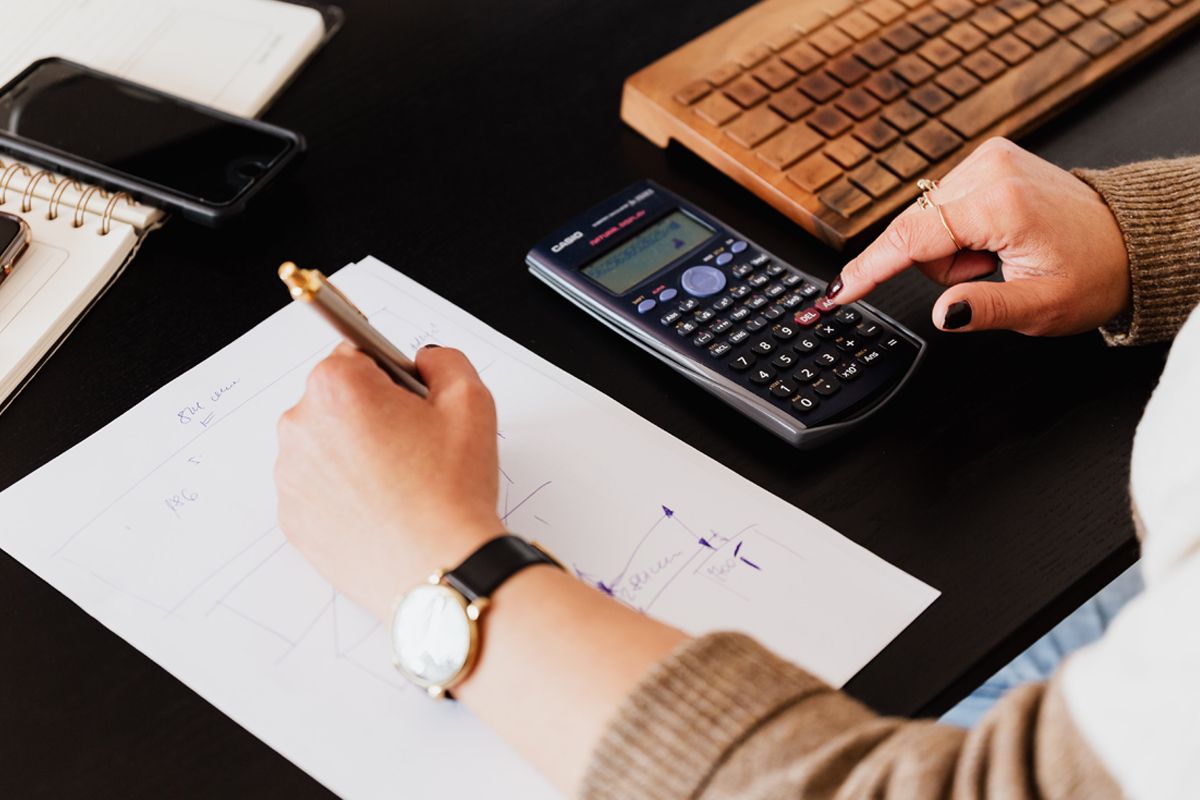 Custom Packaging Cost - Close-up of woman’s hands on a desk making a sketch on a piece of paper with one hand, and using a calculator with another hand.