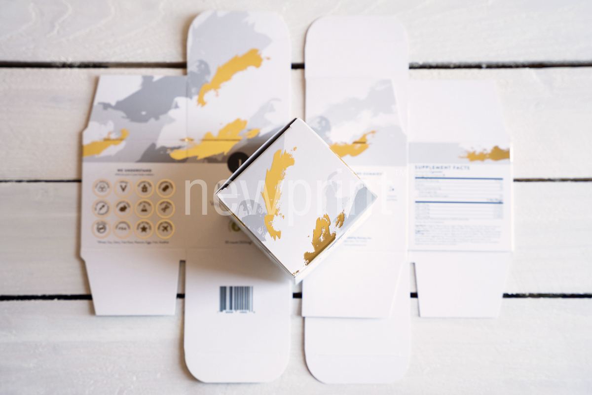 One assembled product packaging box with gold foil details placed on top of the same looking box which is laid out flat on a table.