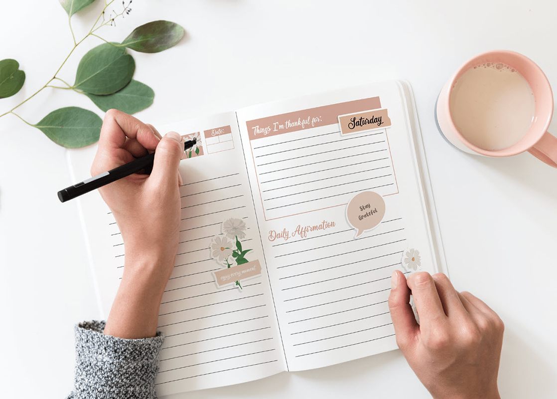 Opened planner on a white table with two hands on top of a planner and one hand holding a pen, custom planner printing.