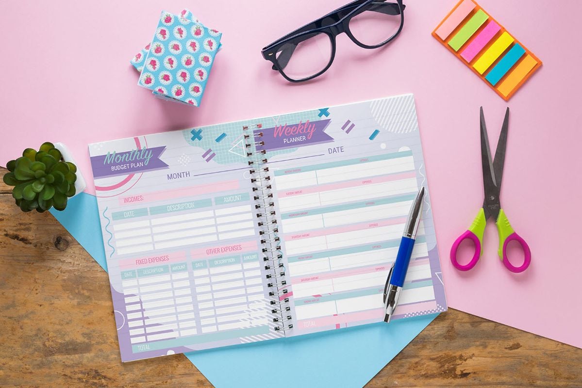 An opened wire bound planner on a wooden desk with a blue and pink paper sheet under the planner, a pen on top of the planner, and stickers, glasses and scissors next to the planner.